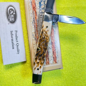 May include: A W.R. Case & Sons folding pocket knife with two silver-colored steel blades and a textured, mottled brown and cream handle. The knife is displayed on its original rectangular box featuring an American flag design and product information.
