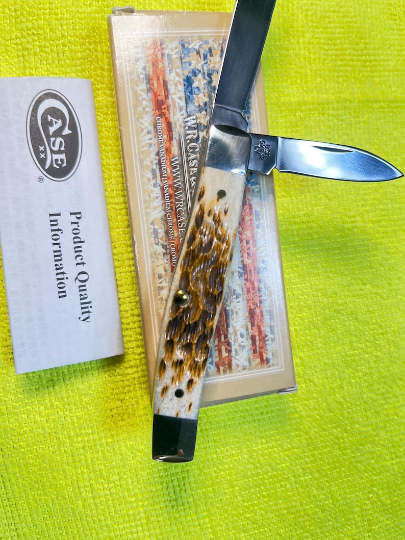 May include: A W.R. Case & Sons folding pocket knife with two silver-colored steel blades and a textured, mottled brown and cream handle. The knife is displayed on its original rectangular box featuring an American flag design and product information.