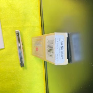 May include: A rectangular beige product box labeled Item No 00077, JACK TEXAS, 62032 CV, and AMBER, featuring a barcode and text. Beside it lies a thin, metallic, cylindrical object encased in a clear plastic sleeve, positioned on a bright yellow cloth.