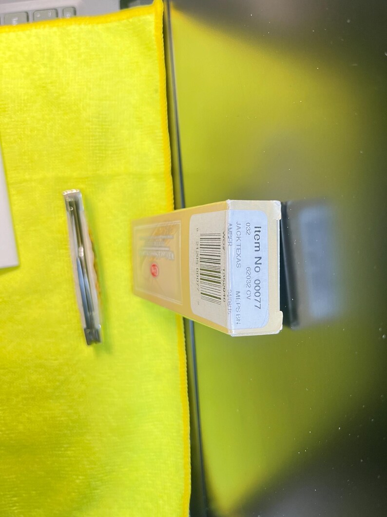 May include: A rectangular beige product box labeled Item No 00077, JACK TEXAS, 62032 CV, and AMBER, featuring a barcode and text. Beside it lies a thin, metallic, cylindrical object encased in a clear plastic sleeve, positioned on a bright yellow cloth.