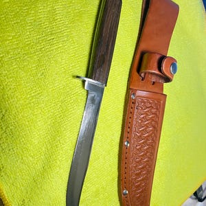Peut inclure: Un couteau de chasse avec un manche en bois et une lame en acier inoxydable. Le couteau est à côté d'un étui en cuir marron avec un motif décoratif et une boucle de ceinture. La lame du couteau mesure environ 20 cm.