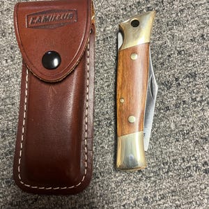 Könnte beinhalten: Eine braune Lederscheide mit dem geprägten Wort "CAMILLUS". Neben der Scheide befindet sich ein geschlossenes Taschenmesser mit einem Holzgriff und Messingakzenten.