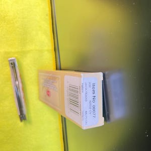 May include: A rectangular tan cardboard box labeled Item No 00077, 62032 CV, JACK TEXAS, and AMBER, featuring a barcode and text. Beside the box lies a slim, metallic folding pocket knife with a light-colored handle, positioned vertically.
