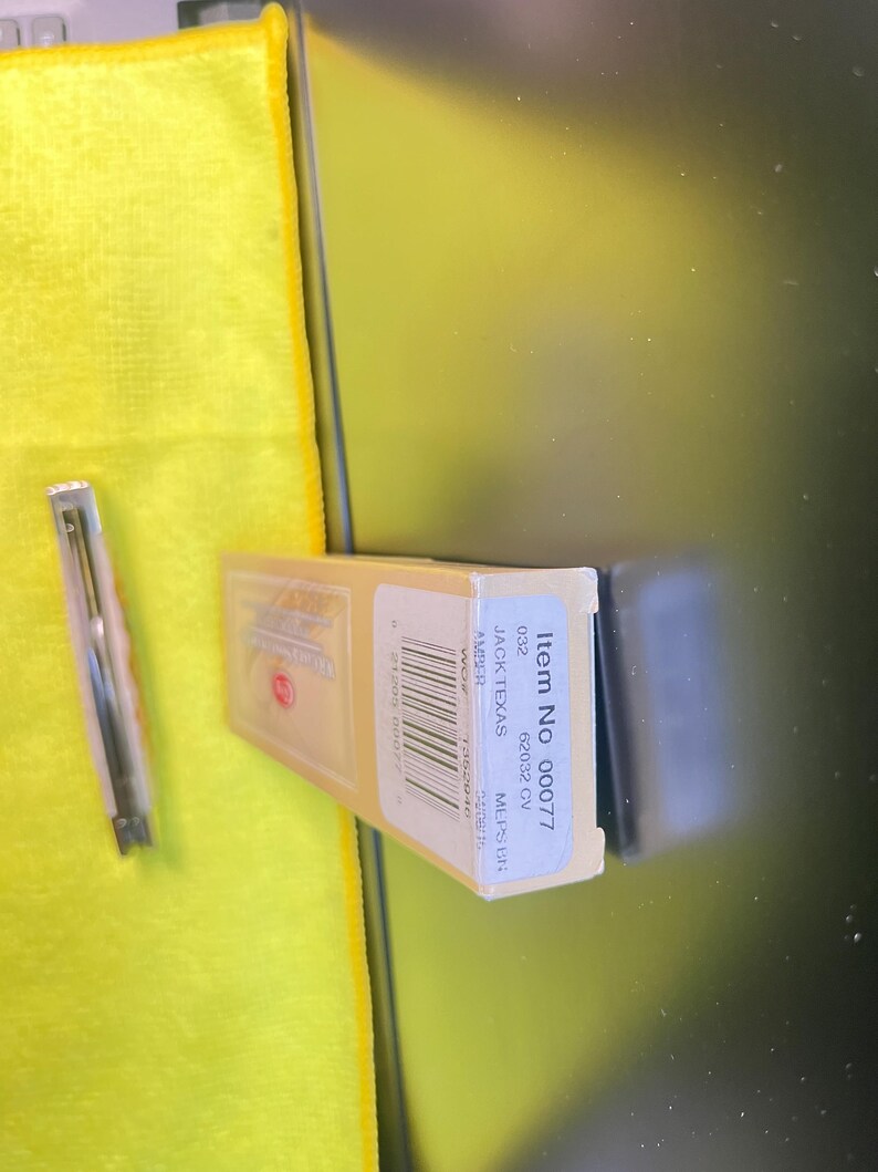 May include: A rectangular tan cardboard box labeled Item No 00077, 62032 CV, JACK TEXAS, and AMBER, featuring a barcode and text. Beside the box lies a slim, metallic folding pocket knife with a light-colored handle, positioned vertically.
