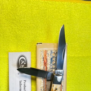 May include: A folding pocket knife with two silver-colored steel blades and a textured, cream and brown handle. The knife is positioned on a tan box featuring a flag pattern and the text W.R. CASE.COM, next to a white paper labeled Product Quality Information.