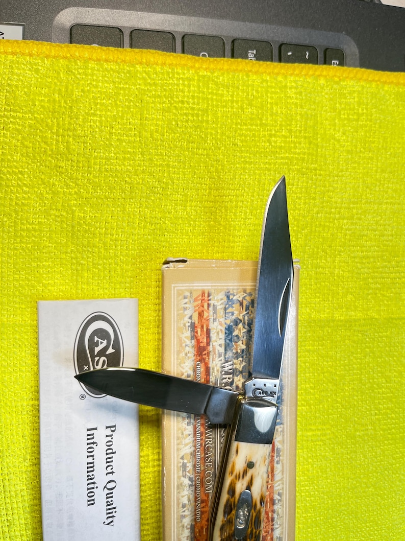 May include: A folding pocket knife with two silver-colored steel blades and a textured, cream and brown handle. The knife is positioned on a tan box featuring a flag pattern and the text W.R. CASE.COM, next to a white paper labeled Product Quality Information.