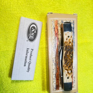 May include: A folding pocket knife with a textured brown and cream handle, featuring silver metal bolsters. The knife is inside a rectangular cardboard box with branding, accompanied by a white paper insert labeled Case Product Quality Information.