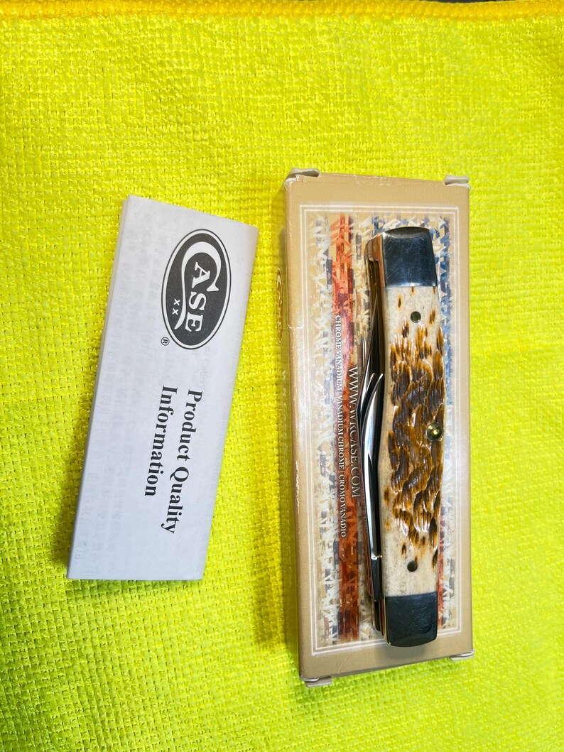 May include: A folding pocket knife with a textured brown and cream handle, featuring silver metal bolsters. The knife is inside a rectangular cardboard box with branding, accompanied by a white paper insert labeled Case Product Quality Information.