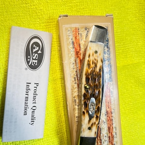 May include: A folding pocket knife with a textured brown and cream handle, featuring a silver bolster and a small metal shield inlay. The knife is packaged in a rectangular box with a flag design, accompanied by a white product information card.