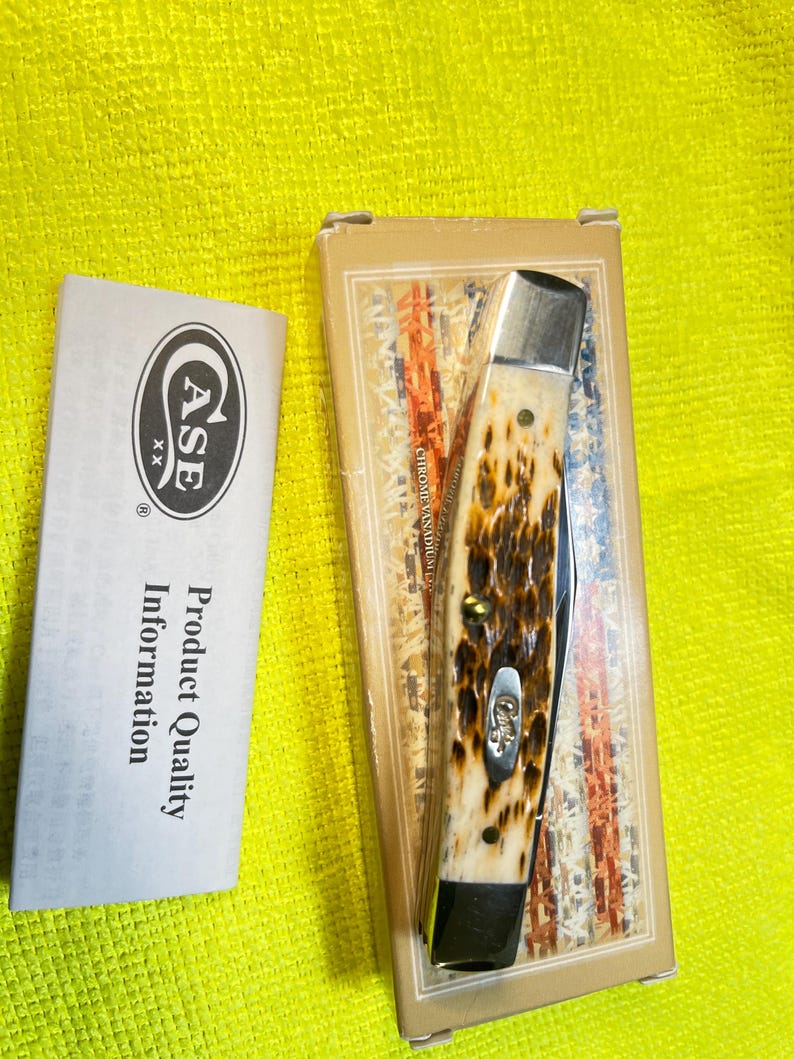 May include: A folding pocket knife with a textured brown and cream handle, featuring a silver bolster and a small metal shield inlay. The knife is packaged in a rectangular box with a flag design, accompanied by a white product information card.
