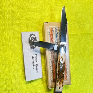 May include: A folding pocket knife with two silver-colored steel blades and a textured brown and cream handle. The knife rests on its rectangular cardboard box, which features a patriotic flag design, alongside a white product information card.