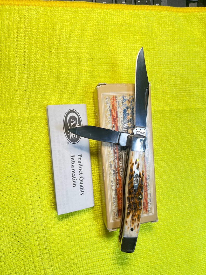 May include: A folding pocket knife with two silver-colored steel blades and a textured brown and cream handle. The knife rests on its rectangular cardboard box, which features a patriotic flag design, alongside a white product information card.