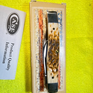 May include: A folding pocket knife with a textured, mottled brown and cream handle and dark metal bolsters, resting on its original rectangular cardboard box. Beside the box is a white paper insert labeled Case XX Product Quality Information.