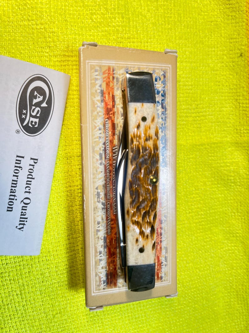 May include: A folding pocket knife with a textured, mottled brown and cream handle and dark metal bolsters, resting on its original rectangular cardboard box. Beside the box is a white paper insert labeled Case XX Product Quality Information.