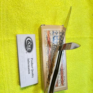 May include: A folding pocket knife with a textured bone handle and two polished metal blades, one partially open. It is positioned next to its rectangular cardboard box featuring a flag design and a white paper insert labeled Product Quality Information.