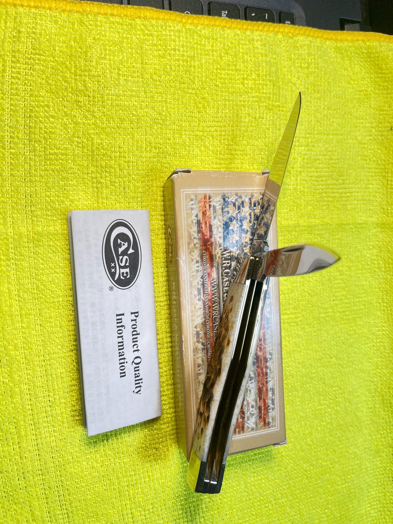 May include: A folding pocket knife with a textured bone handle and two polished metal blades, one partially open. It is positioned next to its rectangular cardboard box featuring a flag design and a white paper insert labeled Product Quality Information.