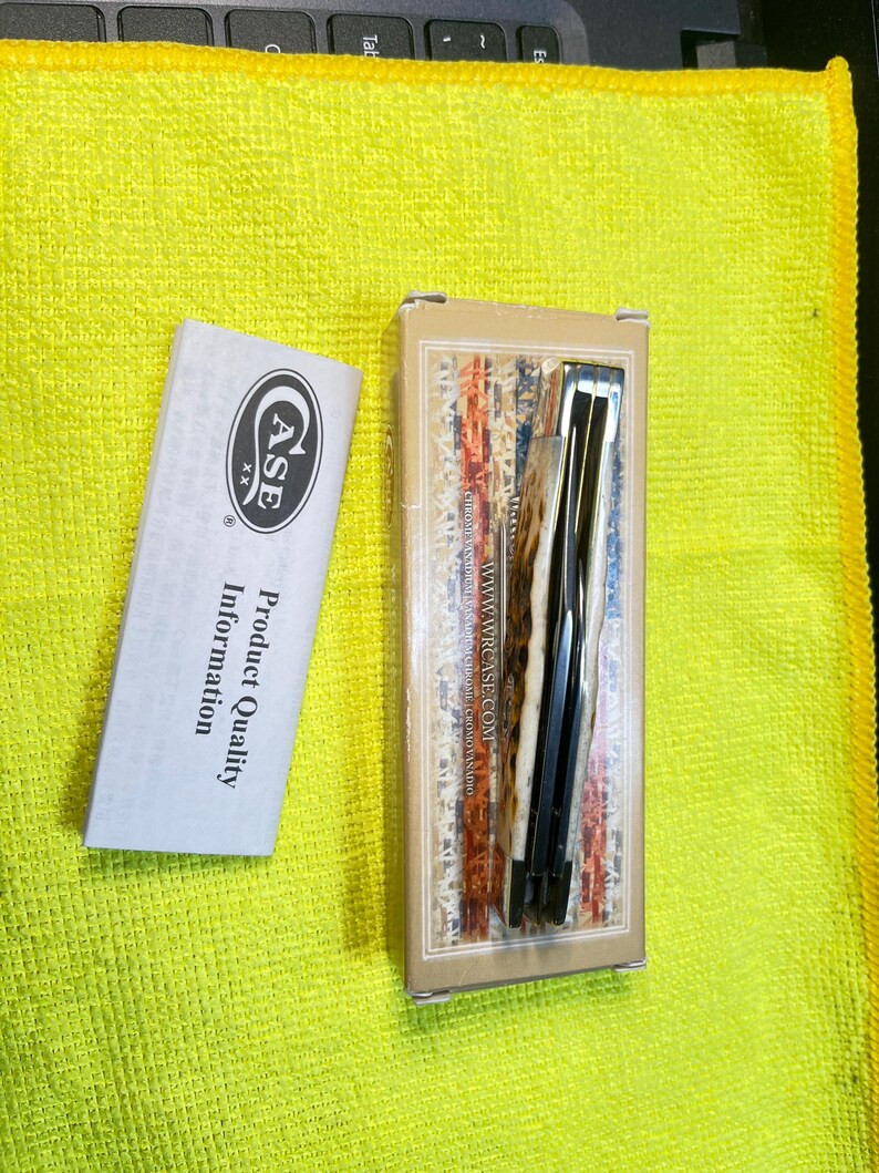 May include: A folding pocket knife in a rectangular tan box with a red, white, and blue flag pattern, featuring the text WWW.WRCASE.COM. Beside the box is a white informational pamphlet with the Case XX logo and the text Product Quality Information.