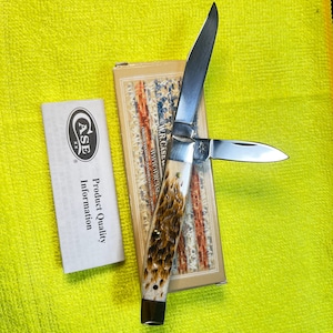 May include: A folding pocket knife with two silver metal blades and a textured brown and cream handle. The knife is positioned on its rectangular cardboard box featuring a red, white, and blue pattern, accompanied by a white product information card.