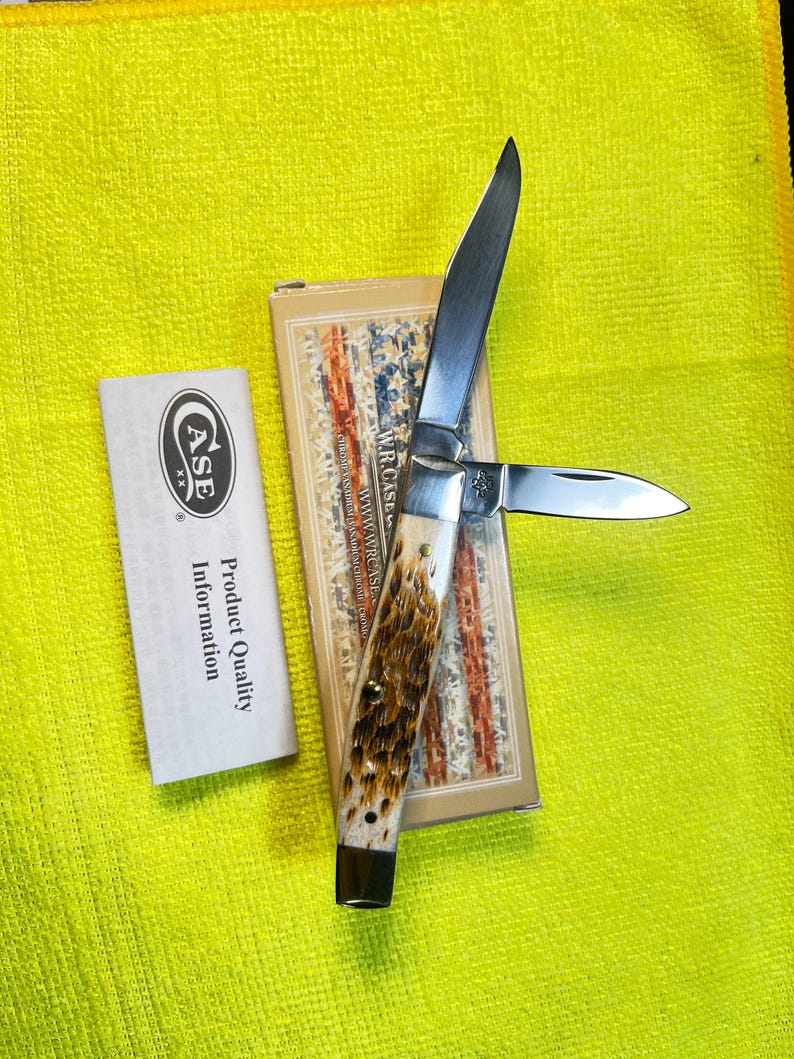 May include: A folding pocket knife with two silver metal blades and a textured brown and cream handle. The knife is positioned on its rectangular cardboard box featuring a red, white, and blue pattern, accompanied by a white product information card.