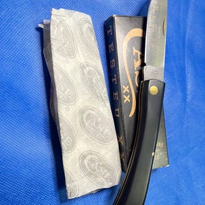 Könnte beinhalten: Ein rostfreies Taschenmesser mit schwarzem Griff und dekorativer Klinge, neben einer Schachtel und einer Papierverpackung. Die Schachtel ist schwarz mit goldfarbenen Buchstaben und die Papierverpackung hat ein sich wiederholendes Muster des Wortes "CASE".