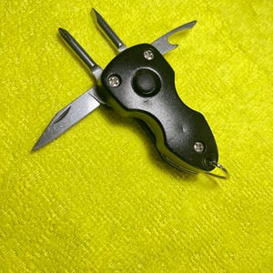 May include: A black multi-tool with several stainless steel attachments, including a knife blade, screwdrivers, and a bottle opener. The tool is open, displaying its various functions. The tool is on a yellow textured surface.