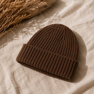 Peut inclure: Un bonnet marron foncé uni doté d'une texture en tricot côtelé épais. Le bonnet présente un large revers plié à la base et une calotte arrondie, mettant en valeur un motif de points verticaux cohérent sur toute la conception du vêtement.