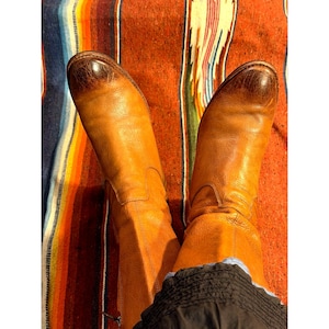 May include: A pair of tan leather boots with a distressed finish and rounded toes. The boots feature a smooth, worn-in texture with darker brown shading at the toe caps and a visible side zipper detail, extending to the mid-calf height.