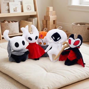 Puede incluir: Una colección de cinco peluches inspirados en el juego Hollow Knight. Las figuras incluyen un caballero blanco con capa gris, un personaje blanco con vestido rojo, una figura naranja con manchas blancas, una criatura blanca y una figura roja y negra.