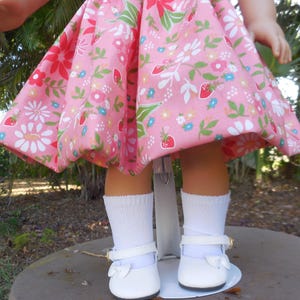 May include: A doll wearing a pink dress with a floral and strawberry pattern. The doll is also wearing white socks and white shoes with a bow and a gold buckle.