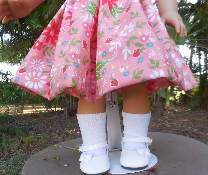 May include: A doll wearing a pink dress with a floral and strawberry pattern. The doll is also wearing white socks and white shoes with a bow and a gold buckle.