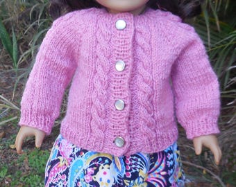 May include: A doll wearing a pink knitted beret and matching cardigan with silver buttons. The doll is also wearing a colorful paisley print skirt, white socks with lace trim, and black shoes. The doll is standing on a round, light-colored surface.