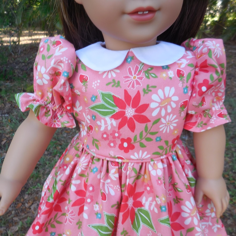 May include: A pink doll dress with a white collar and short, ruffled sleeves. The dress is patterned with red, white, and pink flowers, green leaves, and small strawberries. The dress is designed for a doll.