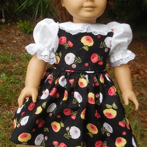 Könnte beinhalten: Eine Puppe mit kastanienbraunem Haar und grünen Augen trägt ein schwarzes Kleid mit einem Blumenmuster aus roten, gelben und weißen Rosen. Das Kleid hat weiße Puffärmel mit Spitzenbesatz, einen weißen Spitzenkragen, dazu trägt die Puppe weiße Socken und schwarze Schuhe.