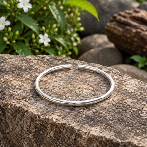 May include: A polished silver-tone open cuff bracelet with a smooth, rounded surface. The bracelet features a simple circular design with a small gap between the two ends, showcasing a reflective metallic finish that catches the light clearly.