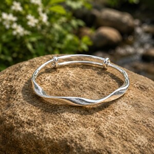 Puede incluir: Una pulsera rígida de tono plateado que presenta una textura ondulada continua a lo largo de su banda. La pulsera está diseñada con un mecanismo deslizante ajustable que consta de dos pequeños bucles cilíndricos que permiten expandir la banda.