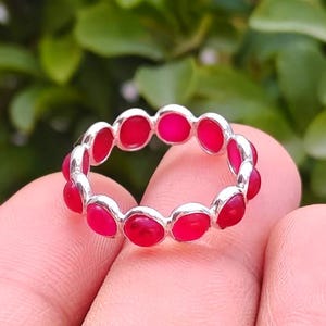 May include: A silver ring featuring a continuous band of small, round, vibrant pink stones set in individual metal bezels. The stones are uniform in size and shape, creating a circular pattern that encircles the finger when worn as a piece of jewellery.
