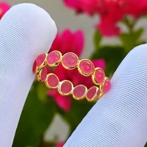Pode incluir: Um anel de banda dourada com uma fila contínua de pequenas pedras cabochão redondas de cor rosa vibrante, engastadas em estilo bisel. O anel é segurado entre dois dedos cobertos por tecido branco, exibindo o metal dourado polido e as pedras.