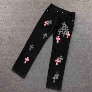 May include: Black denim jeans featuring a straight-leg cut, a button fly, and multiple cross-shaped patches. The patches alternate between solid pink fabric and a black-and-white checkered pattern, distributed across the front of the legs.