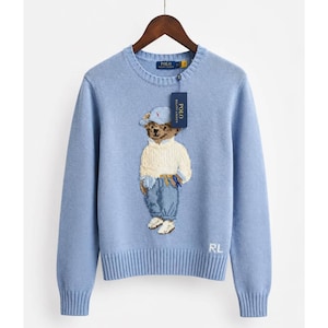 Könnte beinhalten: Ein hellblauer Langarmpullover mit Rundhalsausschnitt und einem Strickmotiv eines Teddybären, der eine Mütze, einen cremefarbenen Pullover und eine blaue Jeans trägt. Der Pullover hat gerippte Bündchen und Saum sowie die in Weiß gestickten Buchstaben RL.