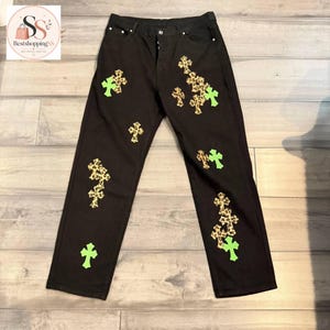 May include: Black denim jeans featuring a button fly closure and a series of scattered cross-shaped patches. The patches alternate between a brown and black leopard print pattern and a solid neon green color, distributed across the front legs.