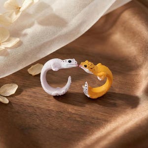 May include: Two open-ended rings shaped like leopard geckos. One ring is white with small black spots and a pink tongue, while the other is yellow with brown spots. Both rings feature textured skin details and curved bodies that form the band.