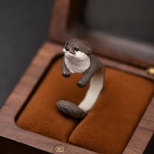 May include: A ring shaped like an otter with a dark brown head and back, a white chest and face, and a textured tail. The otter's body forms the band of the ring, with the head and front paws extending upward and the tail curving at the base.