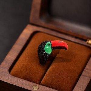May include: A ring shaped like a toucan head with a black textured body, a bright green face patch, and a large, smooth, vibrant red beak. The ring is displayed inside a dark brown wooden box with a soft, brown velvet interior lining.