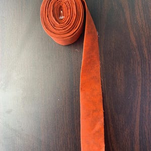 May include: A roll of burnt orange fabric ribbon with a textured, slightly mottled surface. The ribbon is tightly coiled at the top, with a single straight length extending downwards, featuring raw, frayed edges at the bottom end of the strip.