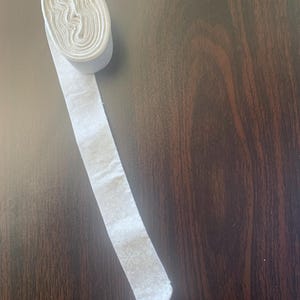 May include: A roll of white fabric ribbon featuring a subtle, repeating snowflake pattern. The ribbon is wound into a tight cylinder, with one end extending outward to display the light-colored textile and the delicate winter-themed print.