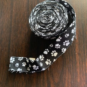 May include: A roll of black fabric ribbon featuring a repeating pattern of white paw prints. The ribbon is coiled into a circular shape with one end extending outward, showing the fabric texture and the consistent white paw print design throughout.