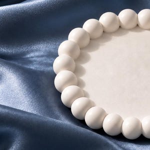 May include: A circular white tray featuring a raised border composed of a continuous row of small, uniform spheres. The tray has a smooth, flat center surface and a matte finish, providing a clean and simple design for holding small items.