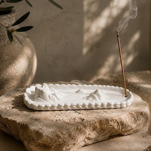 May include: A white ceramic incense holder with a scalloped edge and a sculpted mountain landscape relief on its surface. A single thin brown incense stick stands upright in the holder, emitting a thin trail of white smoke into the air.