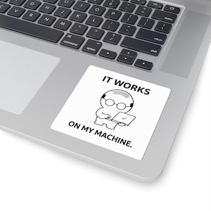 May include: A square white sticker featuring a black line drawing of a bald character wearing glasses and using a laptop. The text IT WORKS ON MY MACHINE is printed in black capital letters above and below the character, with a green checkmark on the screen.