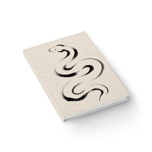 May include: A rectangular hardback journal with a cream-coloured cover featuring a black ink brush stroke illustration of a snake in a winding, S-shaped pose. The cover has a textured appearance, and the notebook is shown from an angled perspective.